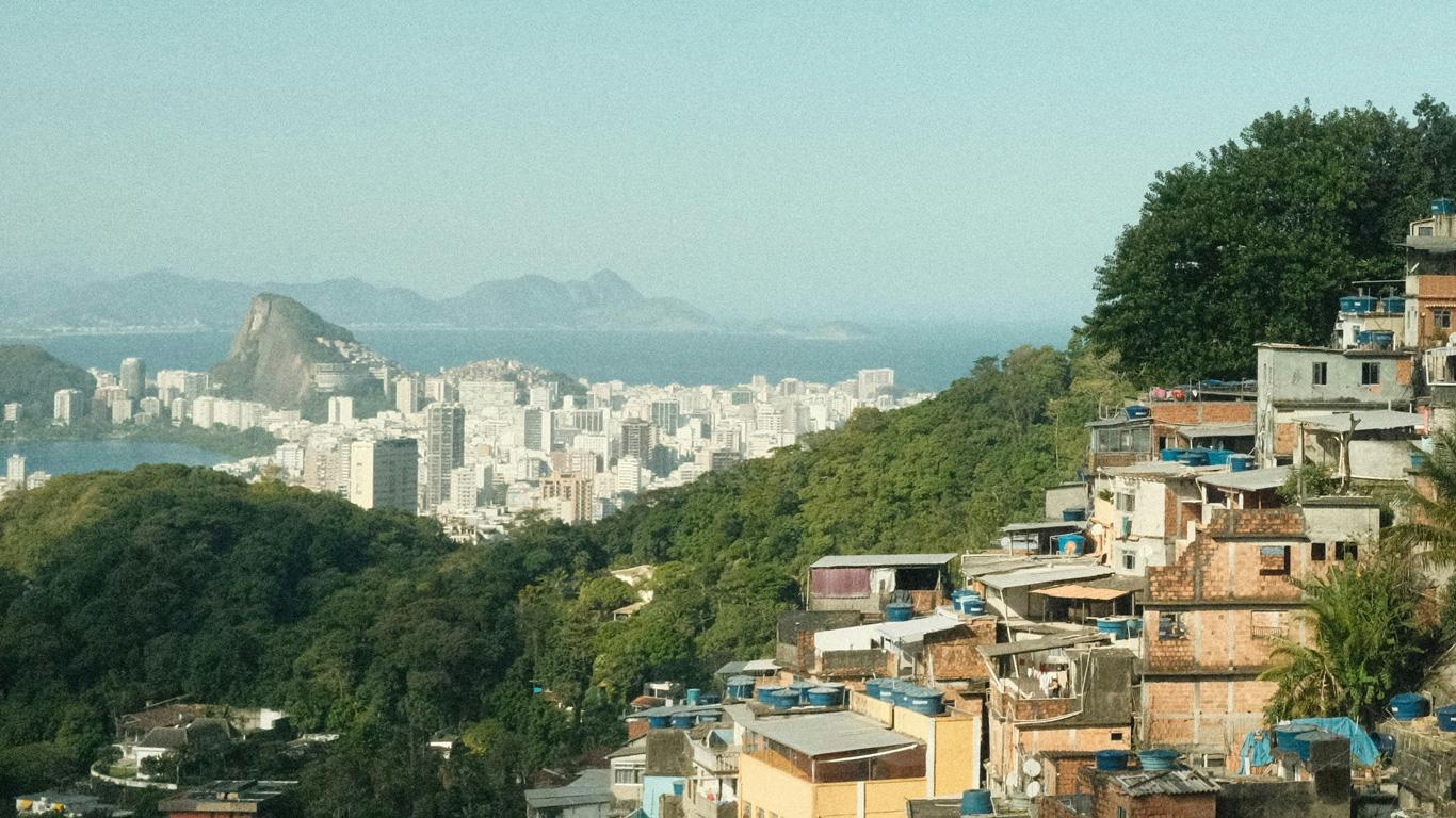 Favela Rocinha en Río de Janeiro con sus casas de colores construidas en terrazas, rodeadas de exuberante vegetación. Al fondo, la ciudad moderna con edificios blancos y el icónico Pan de Azúcar emergiendo en el horizonte junto al mar azul.
