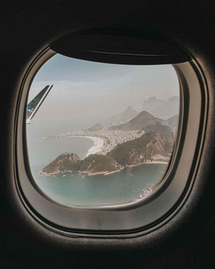 Vista del paisaje costero y las montañas de Río de Janeiro a través de la ventana de un avión, con la punta del ala en la esquina superior izquierda. Ten en cuenta que la hora en Río de Janeiro se encuentra en la Zona Horaria de Brasilia, que es UTC/GMT -3 horas​.