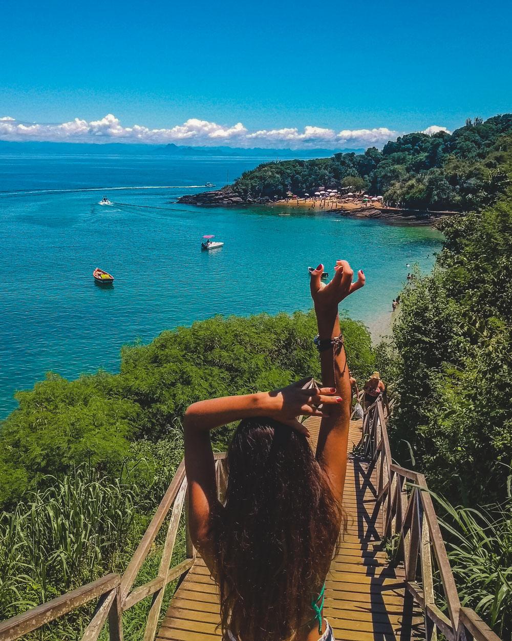 Una mujer, con los brazos levantados en una pose serena, en la Playa de Azeda en Búzios. El paisaje está compuesto por un cielo azul profundo, donde el mar verde se fusiona suavemente en el horizonte. La exuberante vegetación alrededor completa este paisaje encantador.