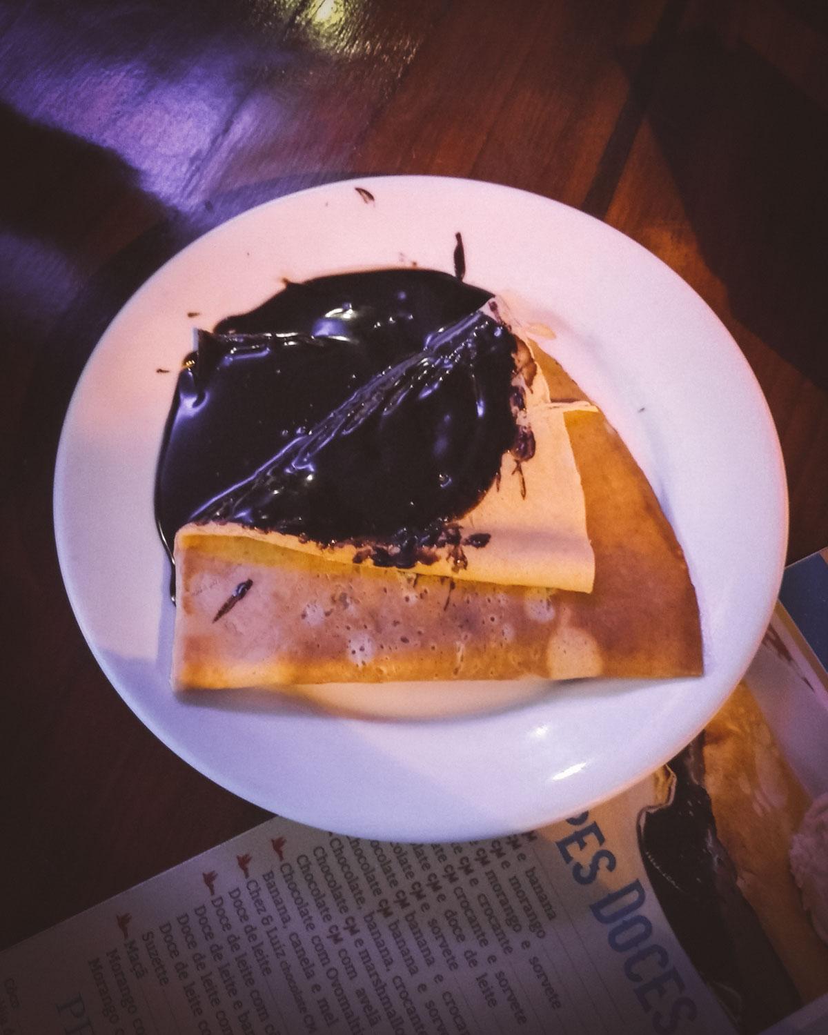 Un crepe cubierto con rica salsa de chocolate, servido en un plato blanco en Chez Michou en Búzios, Brasil, con un menú desenfocado en el fondo.