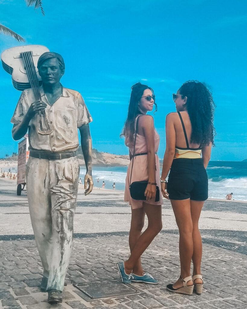 Dos mujeres posando frente a la estatua de Tom Jobim en la Playa de Ipanema. Al fondo, el paisaje está compuesto por el mar azul fundiéndose con el cielo.