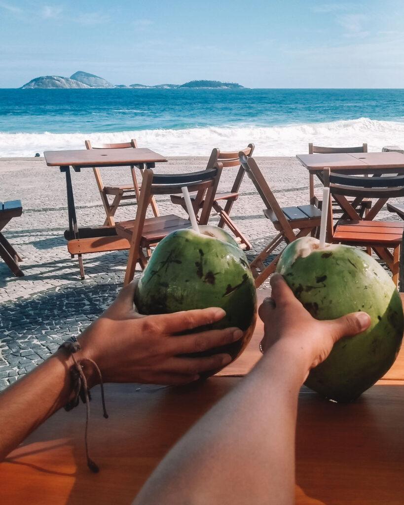 Dos chicas sosteniendo un coco en un chiringuito de la Playa de Ipanema, con el hermoso paisaje de la playa y el mar azul al fondo.