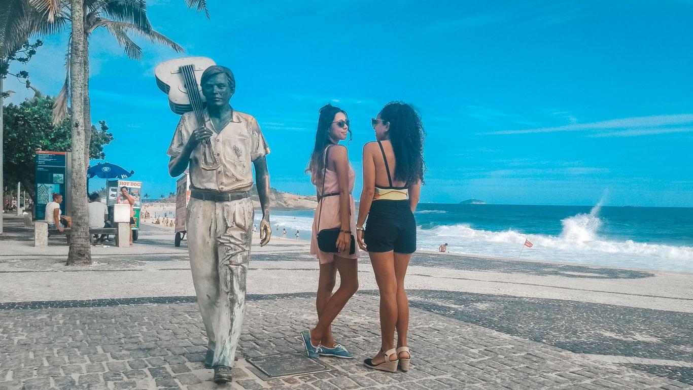 Dos mujeres posando frente a la estatua de Tom Jobim en la Playa de Ipanema. Al fondo, el paisaje está compuesto por palmeras y por el mar azul fundiéndose con el cielo.