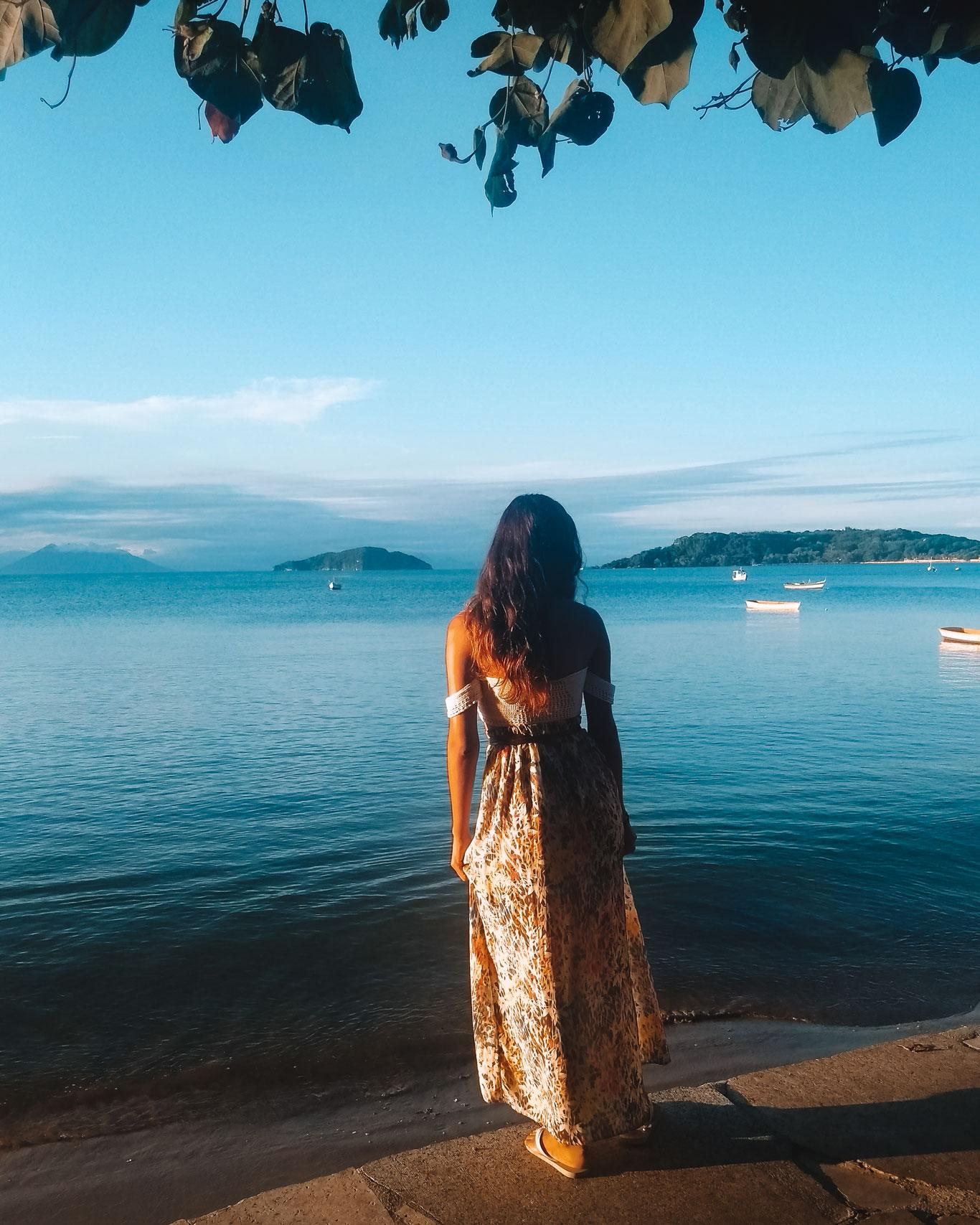 Una mujer de espaldas contempla la serena bahía de Porto da Barra en Búzios, Brasil, vestida con un largo vestido floral. Se aprecia un cielo despejado y un paisaje tranquilo con embarcaciones pequeñas, en contraste con la expectativa de Búzios con lluvia.