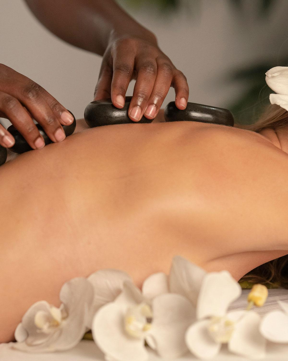 Terapia de masaje en un spa, donde se ven manos realizando el tratamiento sobre la espalda de un cliente relajado, con orquídeas blancas decorativas alrededor.