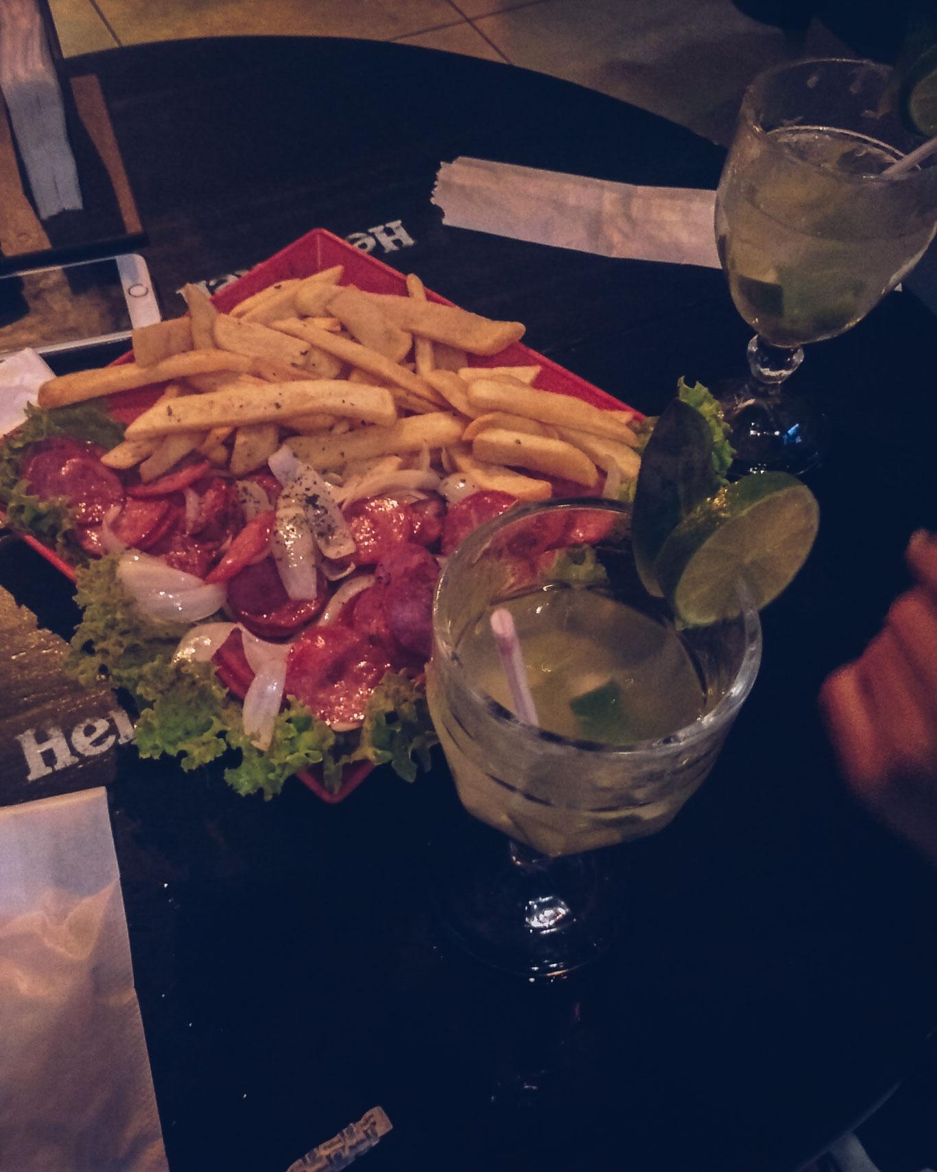 Plato de comida en el bar Zapata en Búzios, Brasil, con una porción generosa de papas fritas y salchichas, acompañadas de dos caipirinhas adornadas con rodajas de lima.