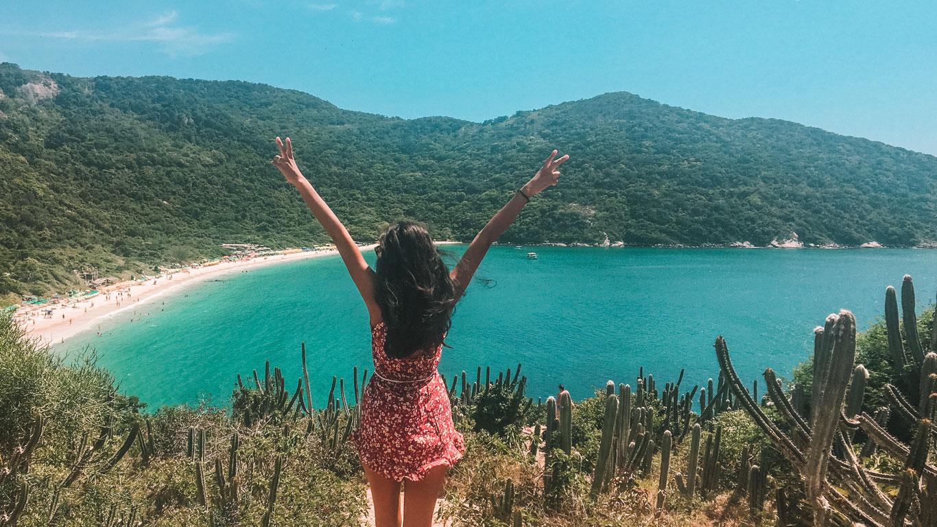 Mujer con los brazos abiertos frente a la Playa do Forno en Arraial do Cabo, Rio de Janeiro, disfrutando de la vista panorámica de la bahía turquesa con montañas al fondo y cactus en primer plano.