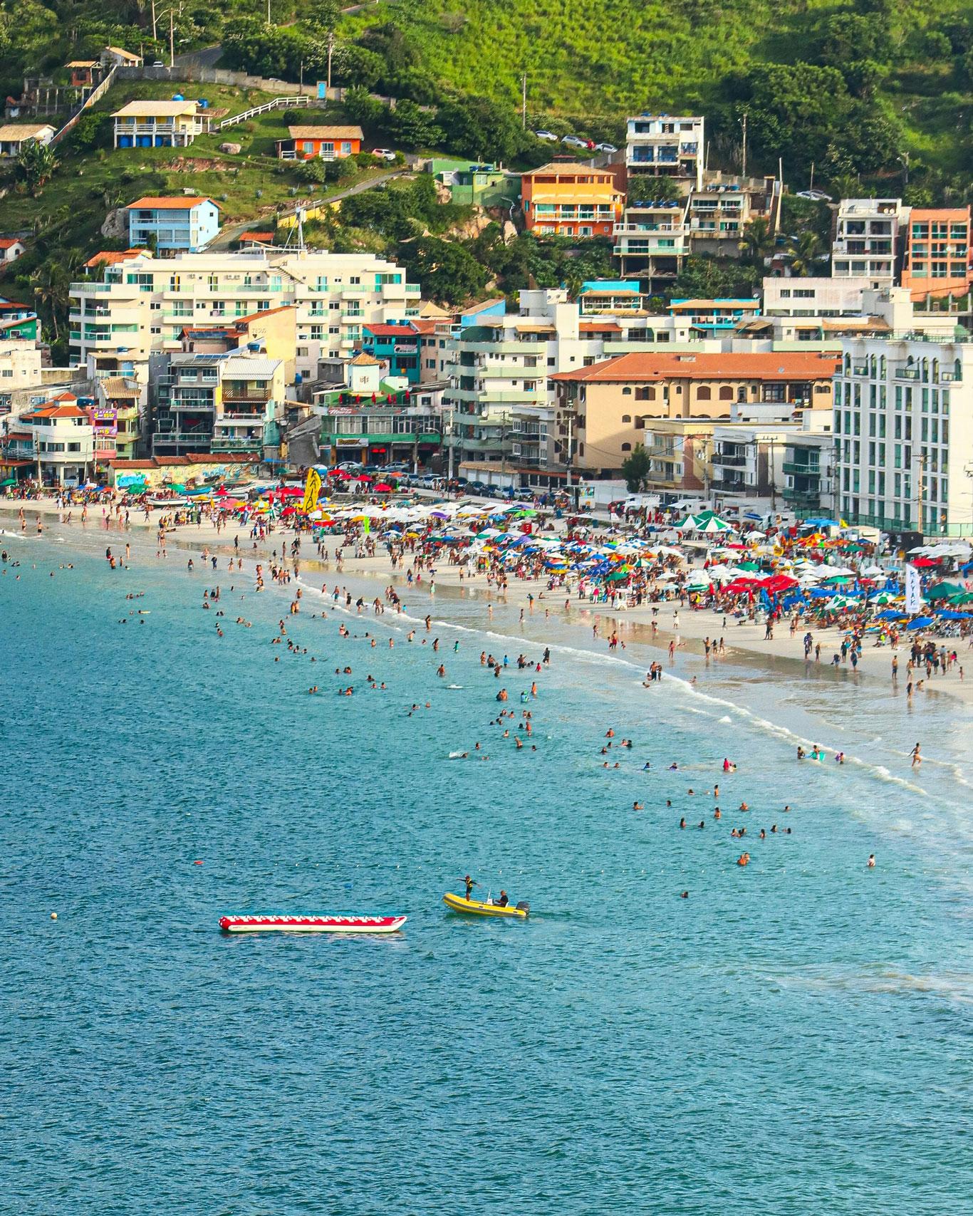 Vista aérea de la Prainha en el verano, una playa concurrida en Arraial do Cabo, con numerosos bañistas y sombrillas coloridas sobre la arena. La playa está rodeada de edificios residenciales de varios pisos y colinas verdes en el fondo, y hay un par de barcos en el agua azul brillante.