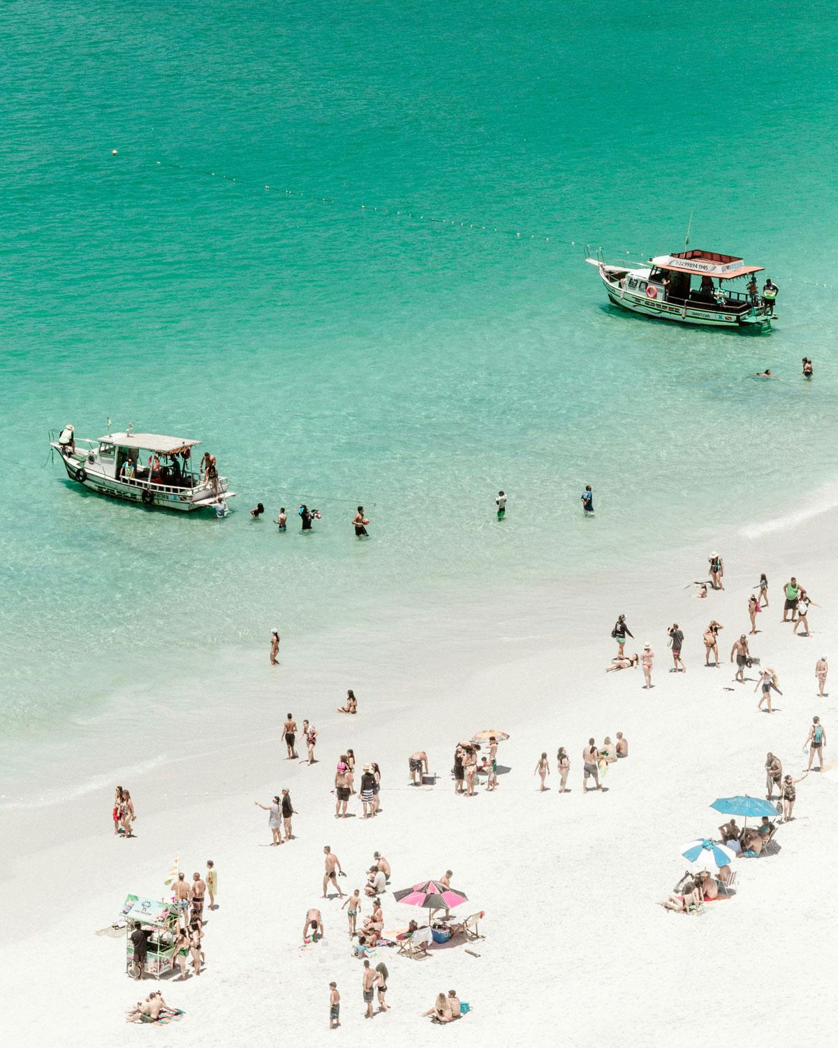 Vista aérea de una animada escena de las Praianhas do Pontal do Atalaia en marzo, la mejor época para ir a Arraial do Cabo, con agua turquesa cristalina, mostrando personas dispersas, bañistas y dos barcos cerca de la orilla.