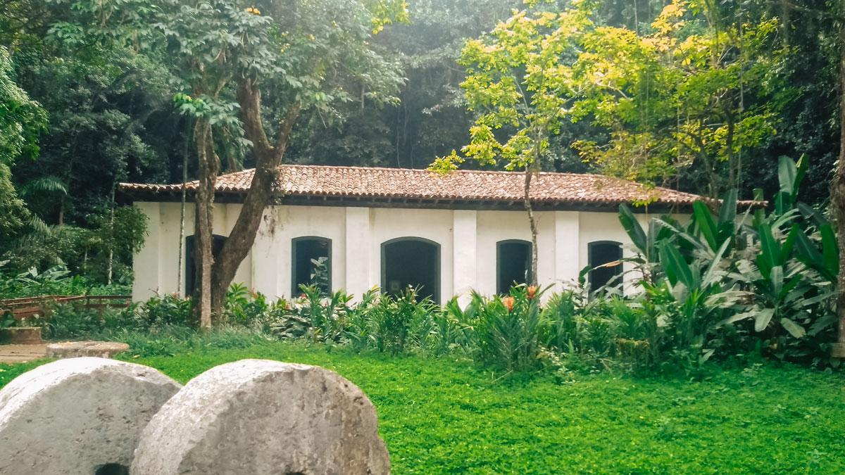 Casa de campo blanca con tejado de tejas rojas rodeada de exuberante vegetación tropical y grandes piedras en el Jardín Botánico de Río de Janeiro, reflejando un ambiente tranquilo y natural.