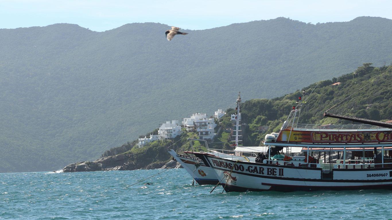 Un barco tradicional amarrado cerca de una costa rocosa con un fondo de colinas verdes y edificios residenciales bajo un cielo despejado, capturado durante un paseo en barco en Arraial do Cabo, en la temporada baja de la ciudad.