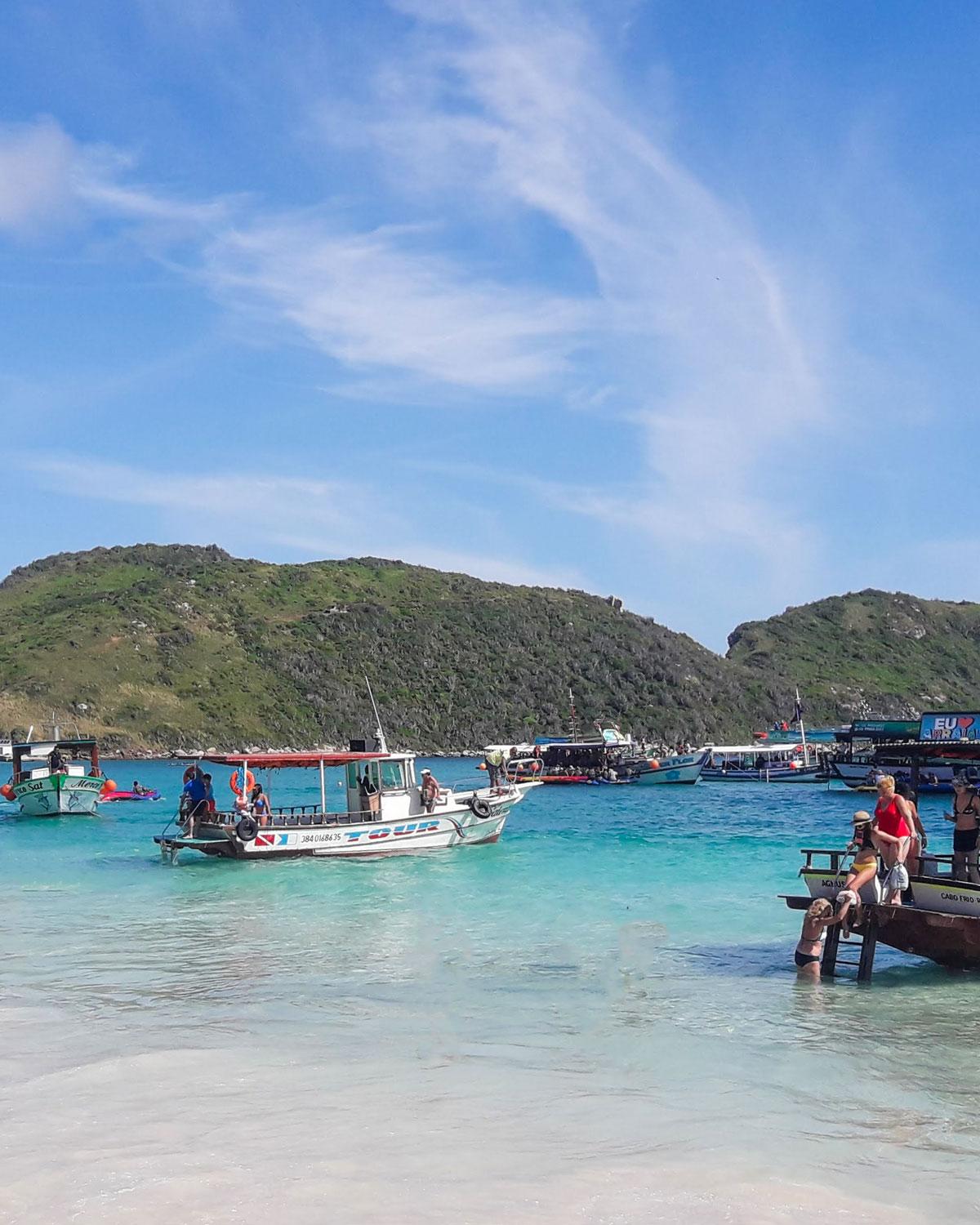 Día animado en la Playa do Farol en Arraial do Cabo, Rio de Janeiro, con visitantes disfrutando de las aguas cristalinas y embarcaciones de turismo ancladas cerca de la orilla, bajo un cielo despejado con una pequeña colina verde al fondo.