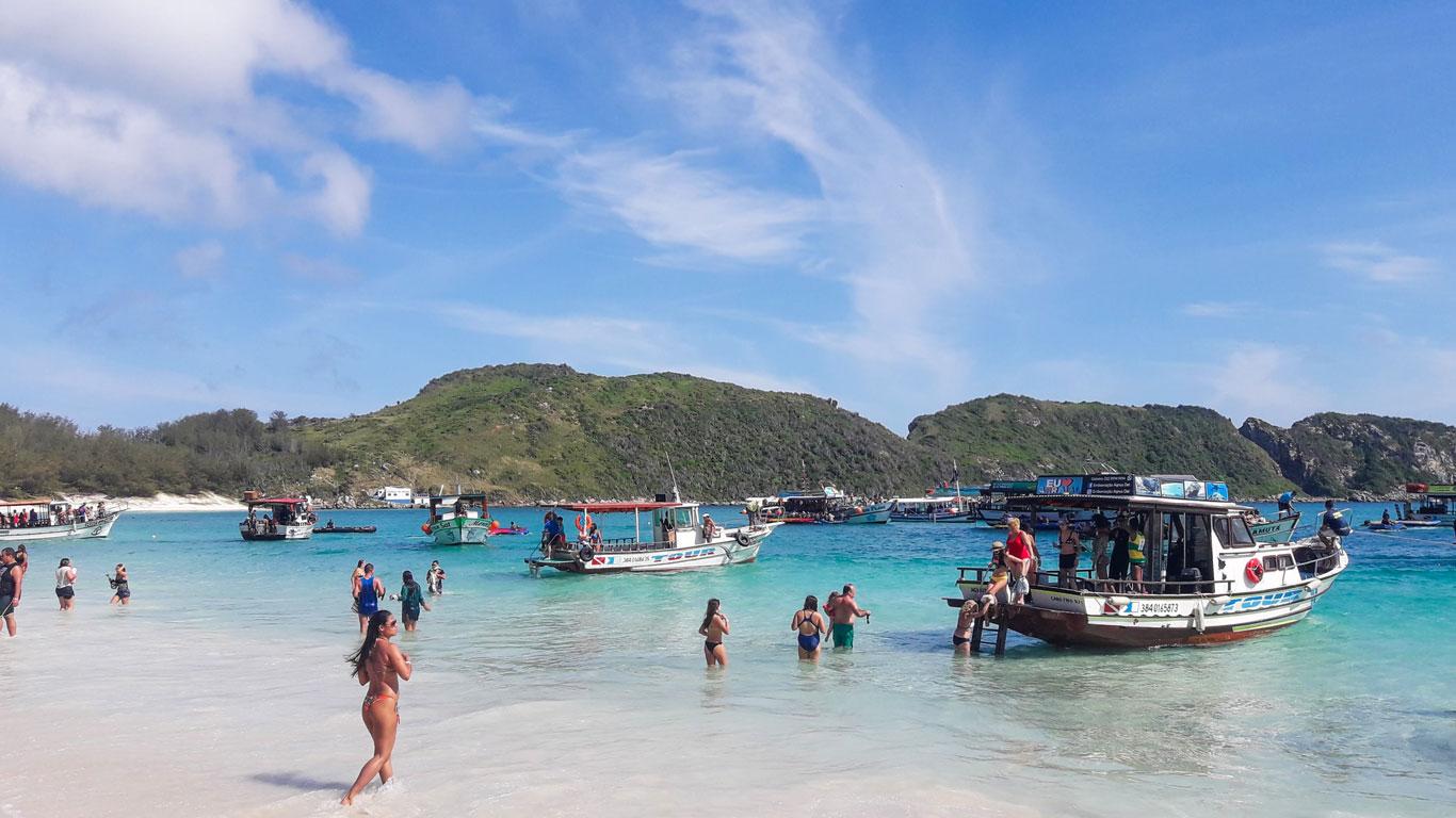 Día animado en la Playa do Farol en Arraial do Cabo, Rio de Janeiro, con visitantes disfrutando de las aguas cristalinas y embarcaciones de turismo ancladas cerca de la orilla, bajo un cielo despejado con una pequeña colina verde al fondo.