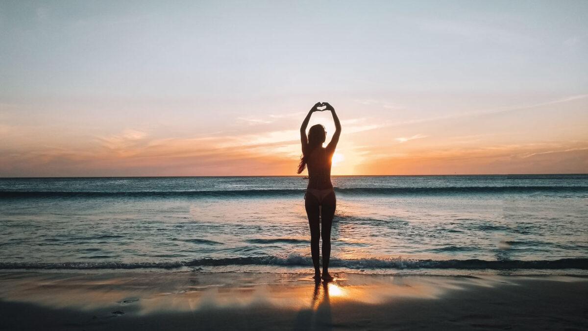 Silueta de una mujer formando un corazón con sus manos sobre su cabeza en la Playa do Grande en Arraial do Cabo, Rio de Janeiro, durante un hermoso atardecer con cielo de tonos cálidos y reflejos dorados sobre las olas suaves.