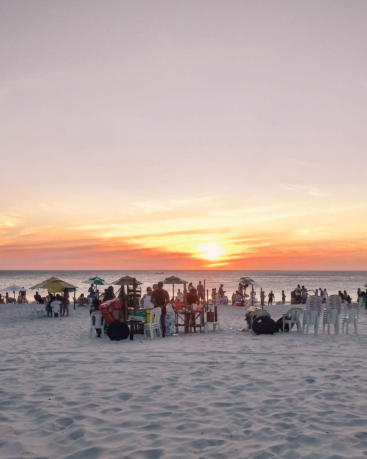 Vista de la Playa do Grande en Arraial do Cabo, Rio de Janeiro, al atardecer con personas disfrutando del ambiente relajado, vendedores en puestos improvisados y el sol descendiendo en el horizonte, tiñendo el cielo de tonos rosados y naranjas.