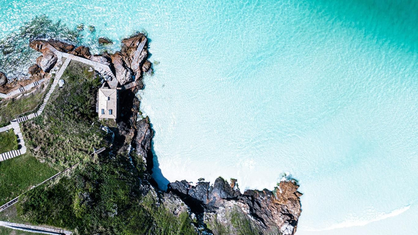 Vista aérea de un tramo rocoso en la Playa do Farol en Arraial do Cabo, con aguas turquesas y una pequeña estructura blanca junto a un camino serpenteante que conecta con la playa.