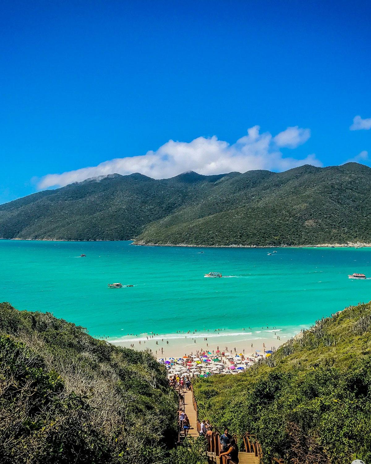 Vista panorámica de las Prainhas do Pontal do Atalaia en Arraial do Cabo, mostrando un sendero descendente rodeado de vegetación, una playa concurrida con sombrillas coloridas y aguas cristalinas, enmarcadas por montañas bajo un cielo azul.