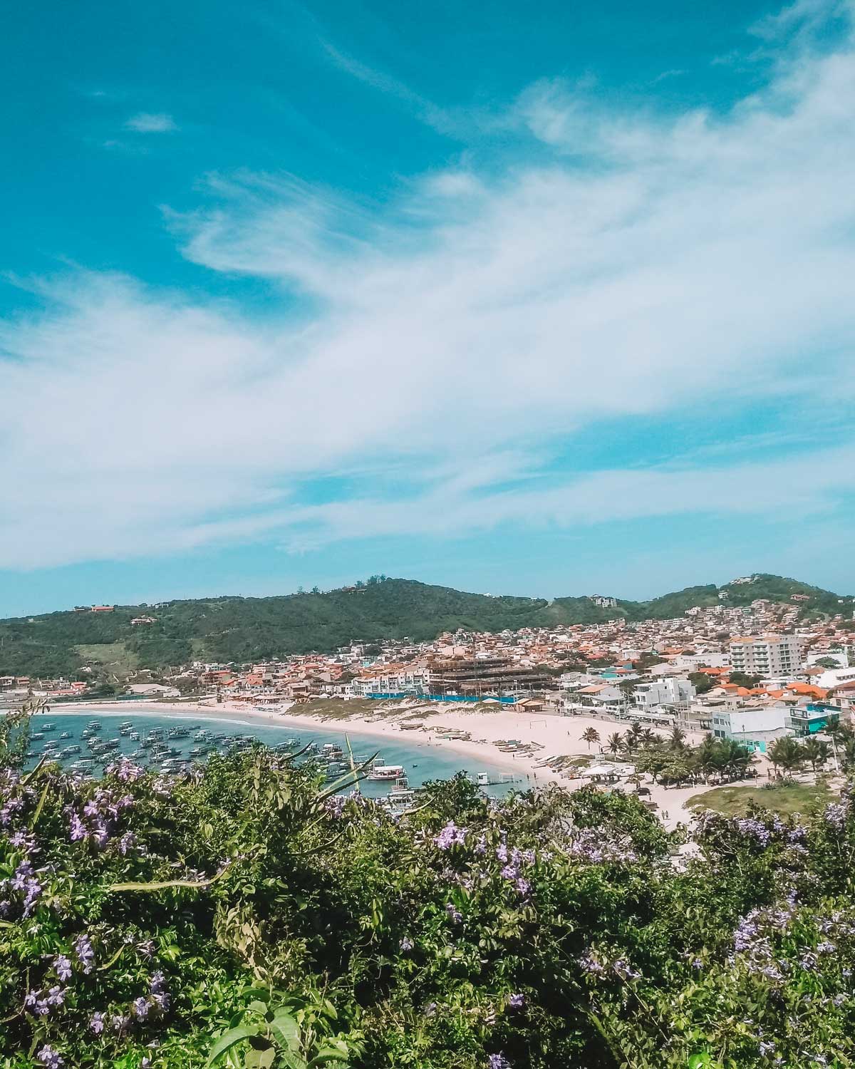 Vista de Arraial do Cabo con una bahía llena de barcos en la Playa dos Anjos, playa de arena y una ciudad que se extiende por las colinas bajo un cielo azul con nubes finas. Flores moradas enmarcan el primer plano de la imagen.