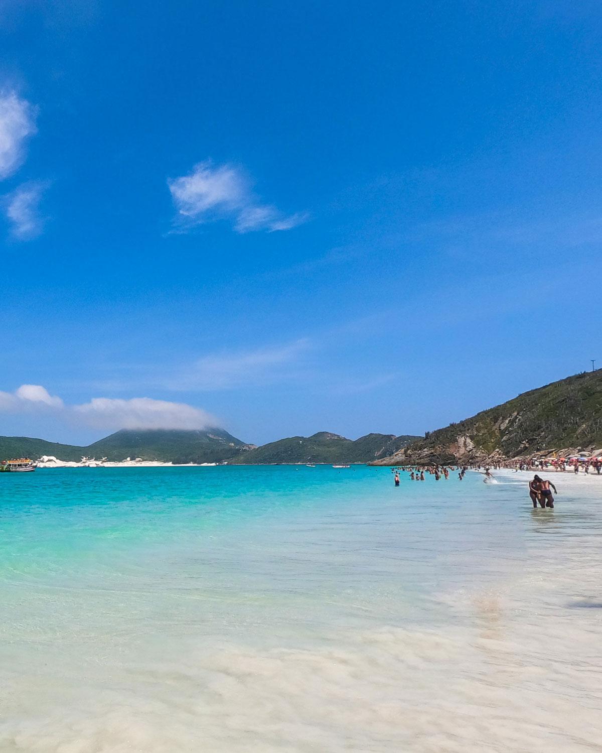 Esta imagen muestra la hermosa Praia do Forno en Arraial do Cabo, destacando las aguas turquesas y cristalinas con suaves olas que llegan a la playa de arena blanca. El fondo presenta montañas verdes frondosas parcialmente ocultas por nubes bajas, bajo un cielo azul vibrante. Se puede ver a personas disfrutando del agua y la playa, añadiendo un ambiente animado al paisaje escénico.