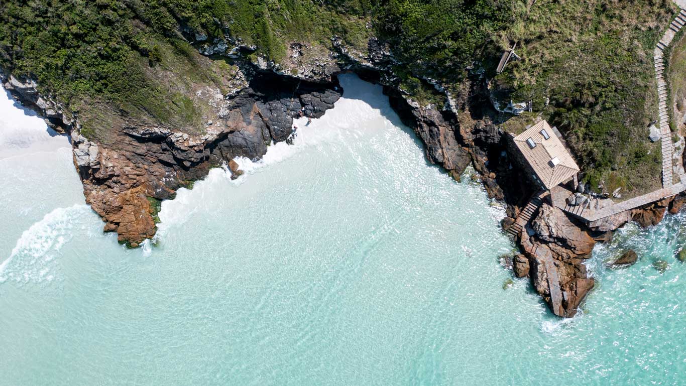 Vista aérea de la costa rocosa con agua turquesa y una pequeña construcción en el acantilado en las Praianhas do Pontal do Atalaia en Arraial do Cabo. Las olas rompen suavemente contra las rocas y un camino conduce hasta la estructura.