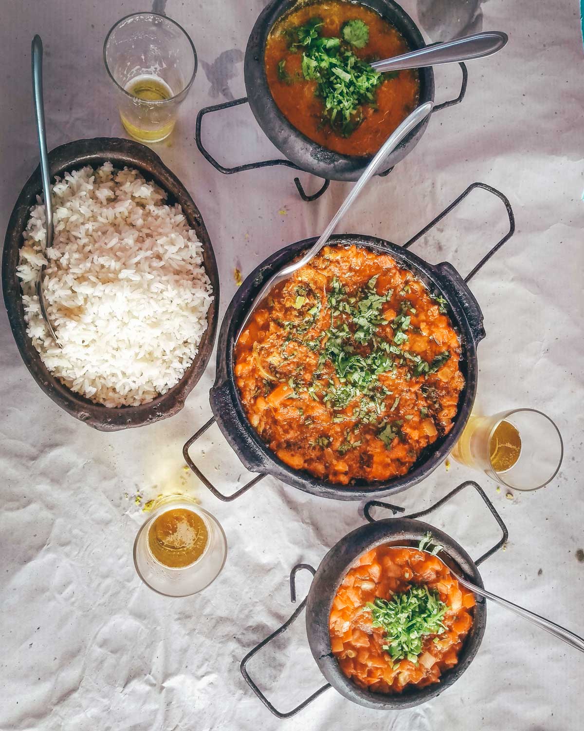 Una mesa servida con moqueca en tres cazuelas de barro, acompañada de un tazón de arroz blanco y vasos de cerveza. Este plato típico brasileño es una delicia que puedes disfrutar en muchos restaurantes de Arraial do Cabo.