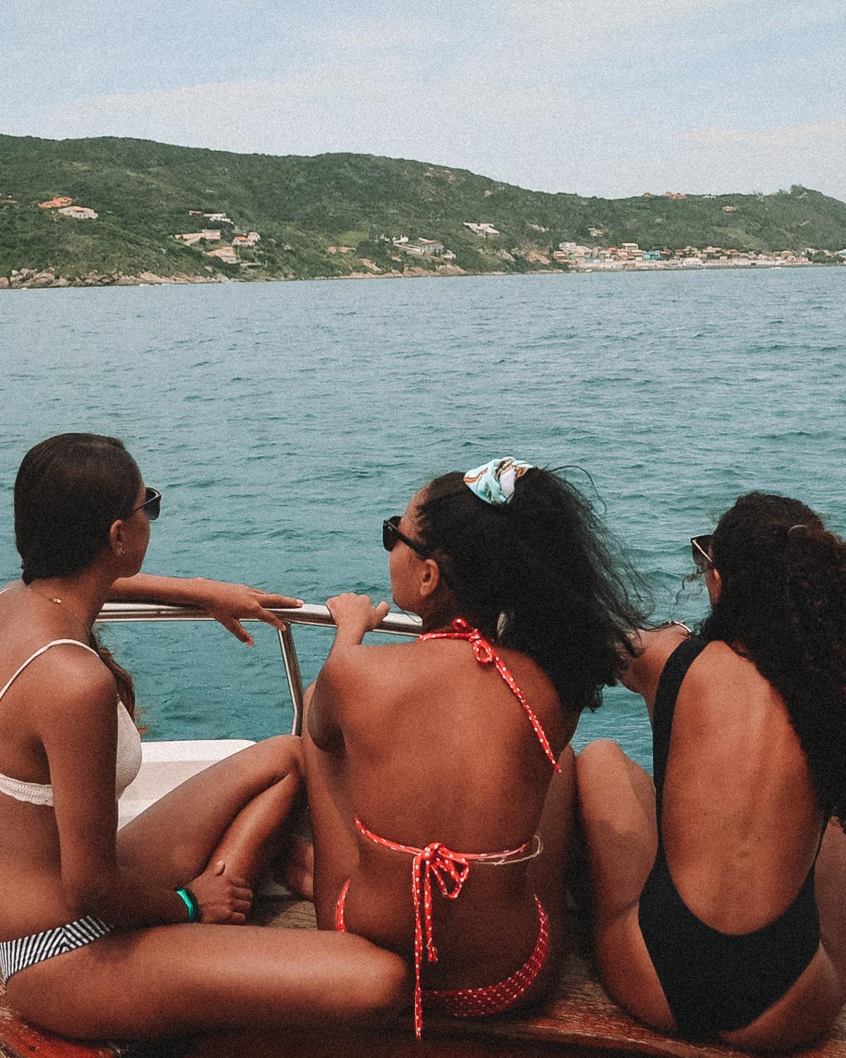 Tres mujeres disfrutando de un paseo en barco en Arraial do Cabo, sentadas en la parte trasera de la embarcación y observando el paisaje costero. Las montañas verdes y las casas dispersas en las colinas se ven en la distancia, reflejando una actividad relajante y escénica en este destino turístico.