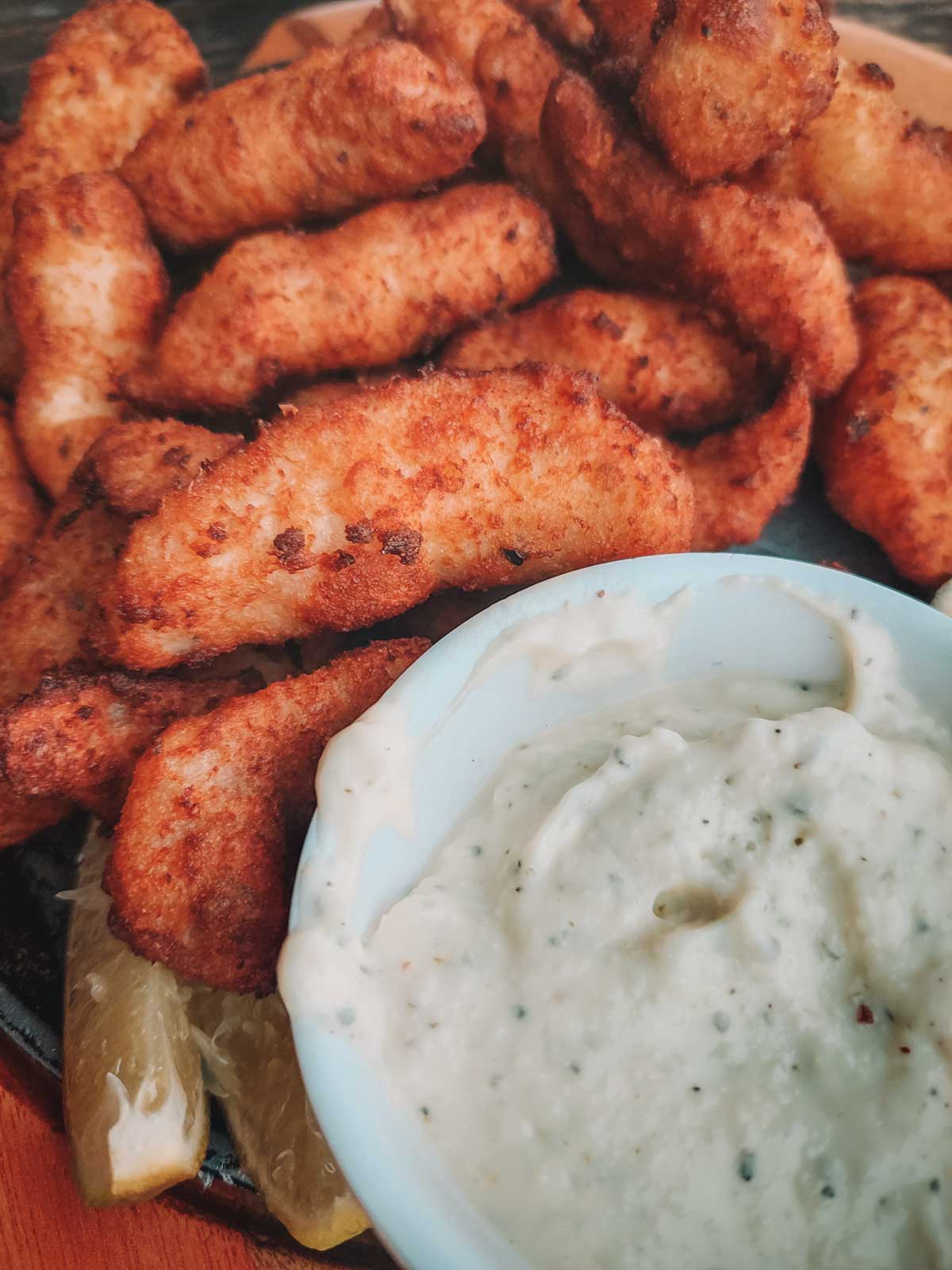 Un plato de iscas de peixe doradas y crujientes servidas en un restaurante de Arraial do Cabo con una salsa cremosa en un tazón blanco, acompañado de rodajas de limón.