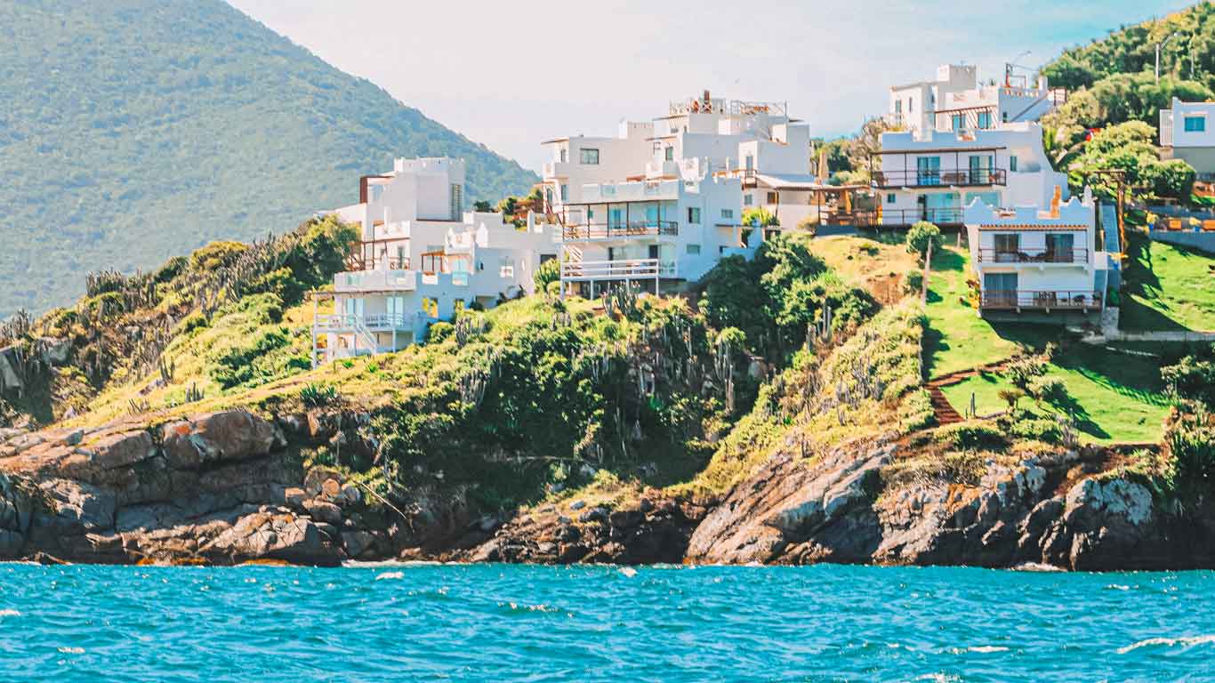 Casas blancas ubicadas en una ladera verde en Pontal do Atalaia,la mejor región para alquilar casa de temporada en Arraial do Cabo, envueltas en niebla con el mar turquesa en primer plano.