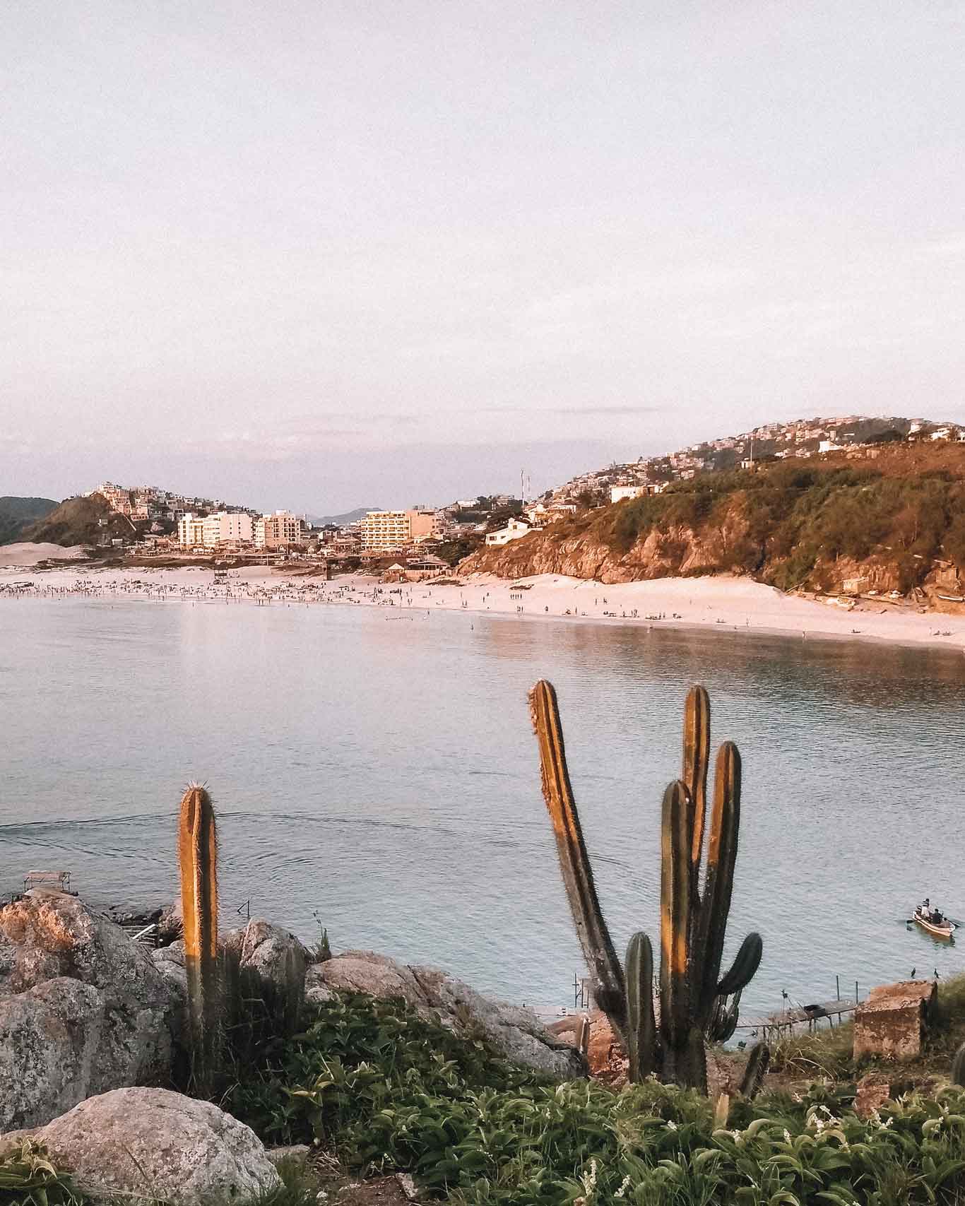 Vista de Playa Grande al atardecer, una de las mejores opciones de playa para alojarse en Arraial do Cabo, con cactus en primer plano y colinas con edificios residenciales al fondo.