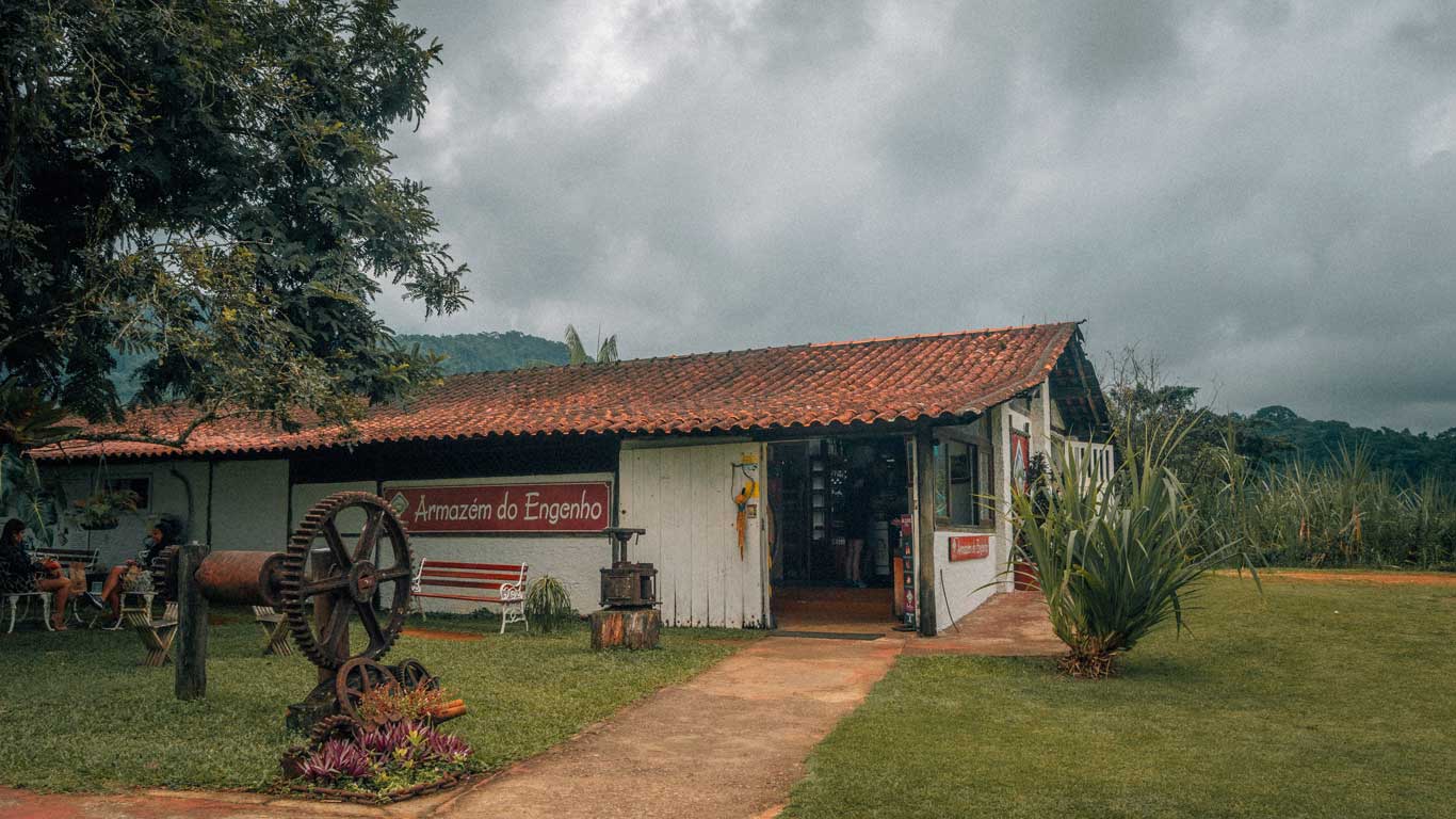 Una tienda rústica con techo de tejas rojas y paredes blancas etiquetada como "Armazém do Engenho", un alambique de Paraty, rodeada de vegetación exuberante bajo un cielo nublado en Paraty, Brasil. Un molino antiguo está expuesto al frente, contribuyendo al encanto de la época.