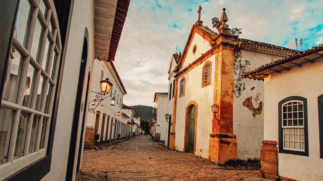 Calle empedrada del Centro Histórico, el mejor lugar donde alojarse en Paraty, al atardecer, con faroles encendidos que iluminan la fachada de una iglesia antigua y edificios coloniales. El ambiente tranquilo y pintoresco resalta la belleza de la arquitectura histórica del lugar.