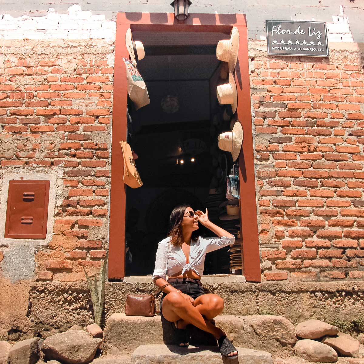 **Texto alternativo en español:** Una mujer sentada en los escalones de una tienda de ladrillos llamada "Flor de Liz" en Paraty. La tienda, especializada en moda de playa, artesanías, y otros artículos, exhibe sombreros y bolsas colgados en la entrada. La mujer, vestida con una blusa blanca anudada y gafas de sol, disfruta del sol mientras mira hacia arriba con una expresión relajada.