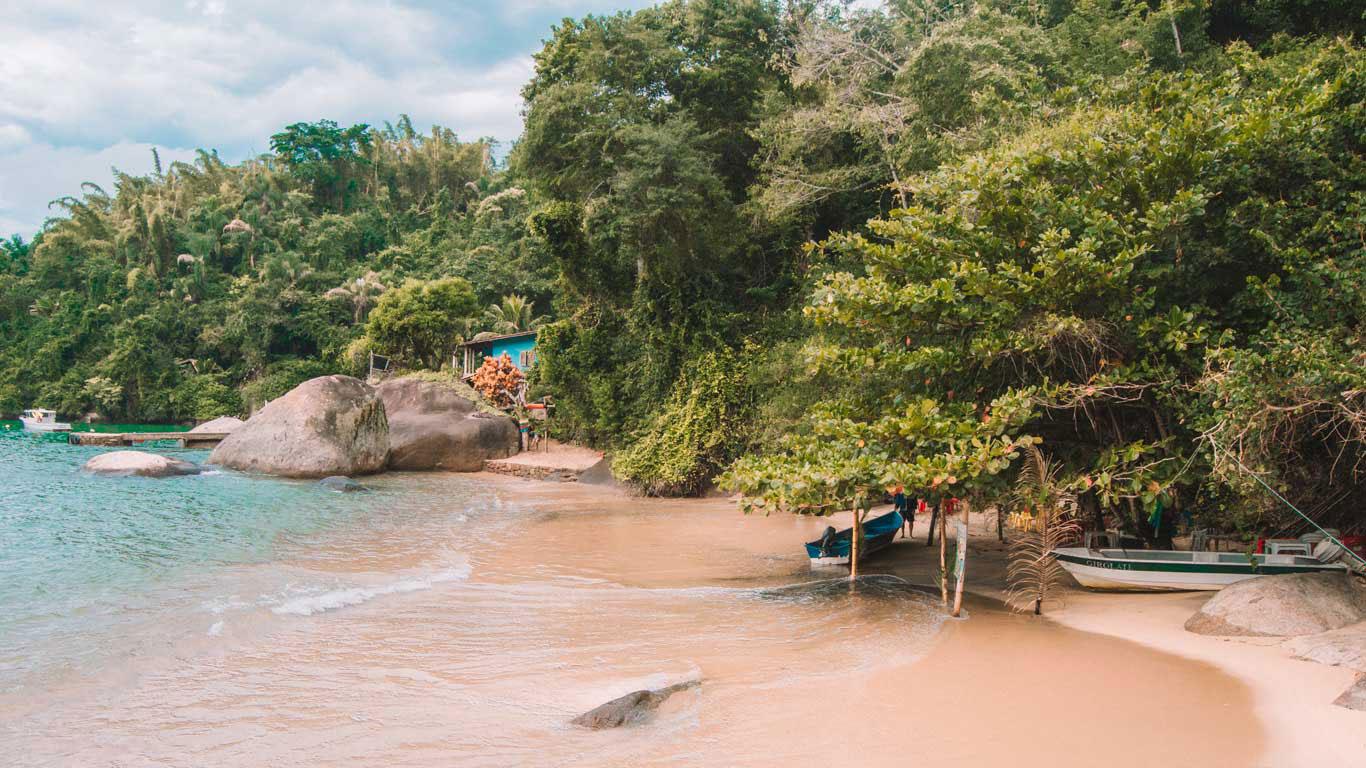 Pequeña playa en Paraty, Río de Janeiro, Brasil, con aguas cristalinas y vegetación densa alrededor. Hay una casa azul y algunas lanchas cerca de la orilla, creando una atmósfera acogedora y serena. Grandes rocas se encuentran dispersas en la arena y el agua, añadiendo al paisaje natural y pintoresco.