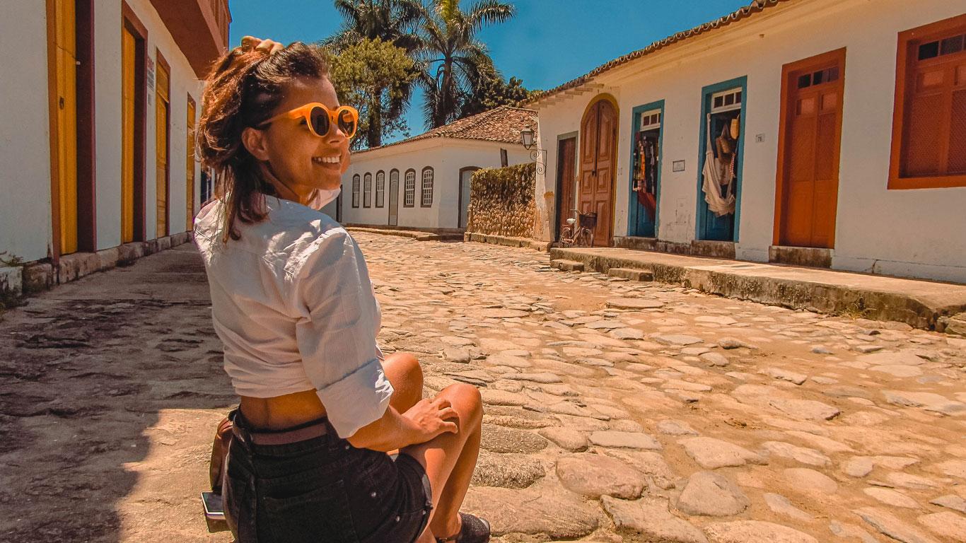 Una mujer con una camisa blanca y gafas de sol naranjas está sentada en una calle adoquinada en el casco histórico de Paraty, Río de Janeiro, sonriendo y disfrutando de un día soleado. La calle está bordeada de coloridos edificios coloniales, con puertas y ventanas pintadas en tonos vibrantes.