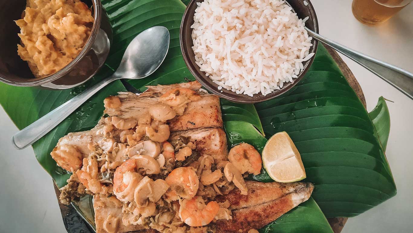 Esta imagen muestra un plato de comida típico de la región de Trindade, en Paraty, Brasil. El plato principal es un pescado con camarones y verduras, servido sobre hojas de plátano que actúan como decoración y plato. Al lado, hay un bol de arroz blanco y otro bol con una guarnición, puré de platano. En el borde del plato, se encuentran dos rodajas de limón. 