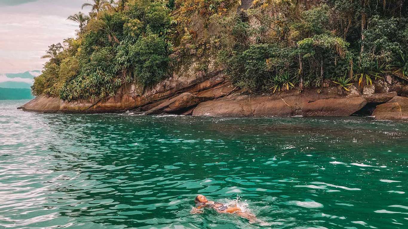 Una mujer nada de espaldas en las aguas cristalinas y turquesas cerca de la Isla dos Cocos en Paraty. Al fondo, se observa una costa rocosa cubierta de densa vegetación tropical, que desciende abruptamente hacia el agua. El cielo está parcialmente nublado, creando un ambiente tranquilo y natural.