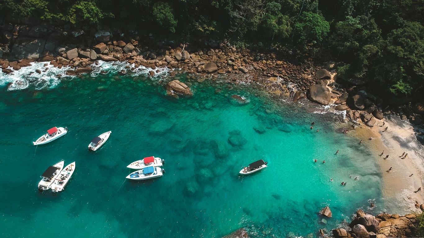 Vista aérea de una playa en Ilha Grande, donde varias lanchas están ancladas en las aguas cristalinas y turquesas cerca de la orilla. Las olas rompen suavemente contra las rocas que bordean la playa, mientras varias personas disfrutan del agua y la arena en este paraíso tropical rodeado de vegetación exuberante.