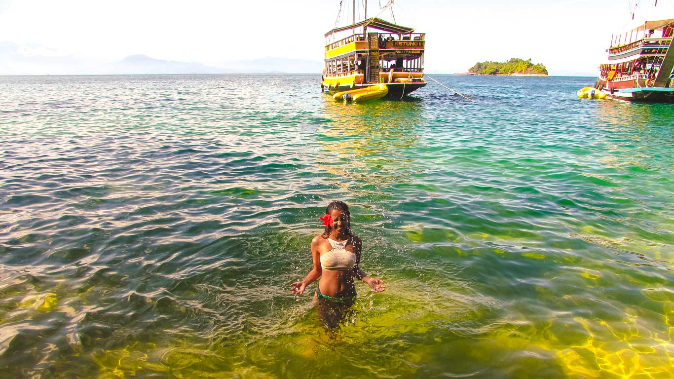 na mujer en bikini, con una flor roja en el cabello, está de pie con el agua hasta la cintura en las aguas turquesas del mar en Paraty. Dos coloridas escunas están ancladas cerca, y se ve una isla en el fondo.