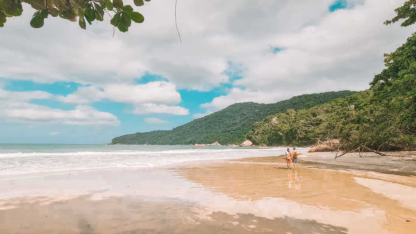 Esta imagen muestra una vista de la Praia do Cachadaço en Trindade, Brasil. La playa es amplia y serena, con arena dorada y olas suaves rompiendo en la orilla. Dos personas caminan a lo largo de la playa, disfrutando del paisaje natural que incluye frondosos árboles a la derecha y colinas verdes al fondo. El cielo está parcialmente nublado, pero se pueden ver áreas de cielo azul, lo que añade un toque vibrante a la escena. Las ramas de un árbol en la parte superior enmarcan la imagen, acentuando la belleza natural del lugar.