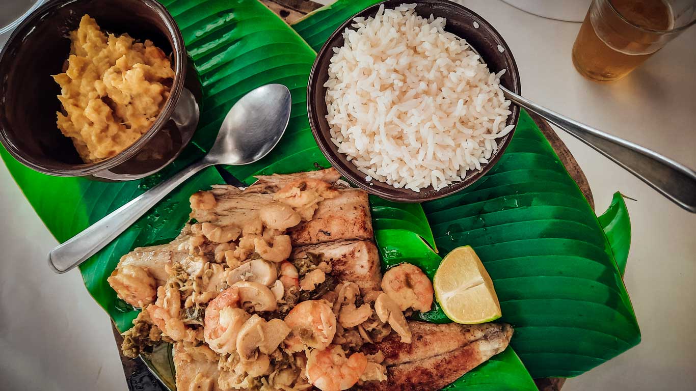 Un plato tradicional brasileño se sirve sobre una hoja de plátano verde, con pescado a la parrilla cubierto de camarones, acompañado de arroz y un acompañamiento. Una rodaja de limón está al lado del pescado, y un vaso de cerveza es parcialmente visible en la mesa.