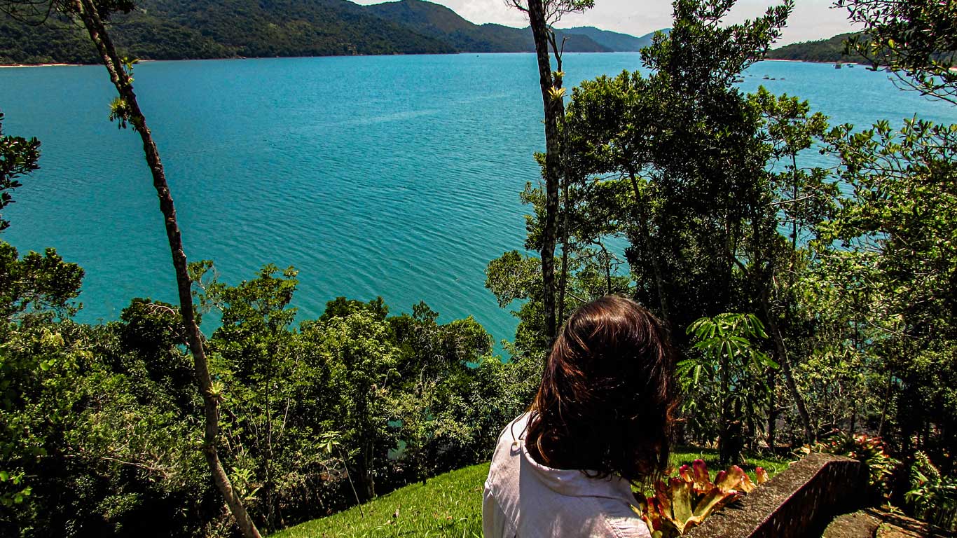 Una mujer mira hacia las impresionantes aguas azules del Saco do Mamanguá, un de los mejores paseos que hacer en Paraty, rodeado de verdes montañas en Paraty. La vista desde el mirador es serena y expansiva, capturando la belleza natural de este brazo de mar.