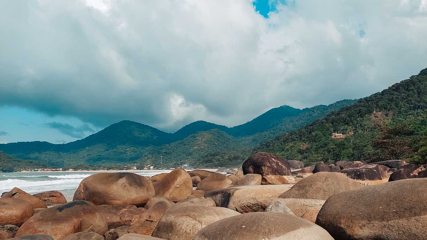 Esta imagen muestra a una persona sentada en una gran roca en la Praia de Fora en Trindade, Brasil. La persona, que lleva un sombrero de tela gris y ropa casual, está mirando hacia un paisaje compuesto de grandes rocas que se extienden a lo largo de la costa, con un fondo de montañas verdes cubiertas de vegetación densa. El cielo está parcialmente nublado, lo que añade una sensación de tranquilidad a la escena. En una de las rocas más grandes, se puede ver un graffiti con el nombre "Thayara". La atmósfera sugiere un momento de reflexión en medio de la naturaleza.