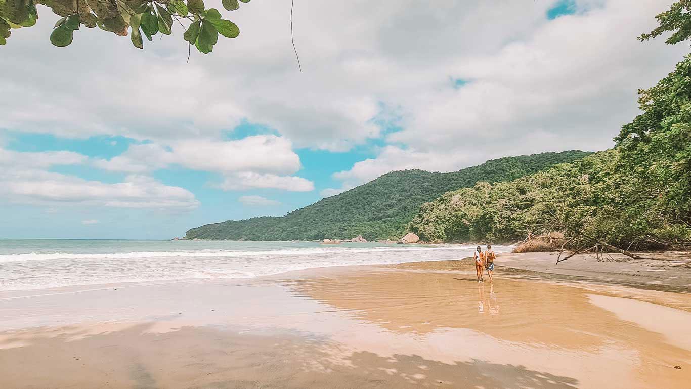 Playa do Cachadaço en Trindade en Paraty, con dos personas caminando por la orilla bajo un cielo parcialmente nublado. La playa está rodeada de exuberante vegetación y colinas, creando un ambiente tranquilo y natural.