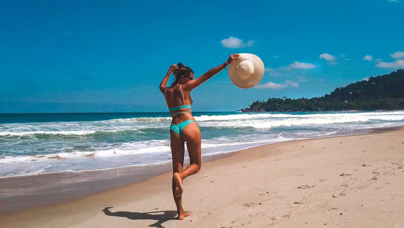 Una mujer en bikini turquesa camina descalza por la arena de una playa en Ubatuba, sosteniendo un sombrero de paja de ala ancha en el aire. Las olas rompen suavemente en la orilla bajo un cielo azul claro, mientras que al fondo se pueden ver colinas cubiertas de vegetación.