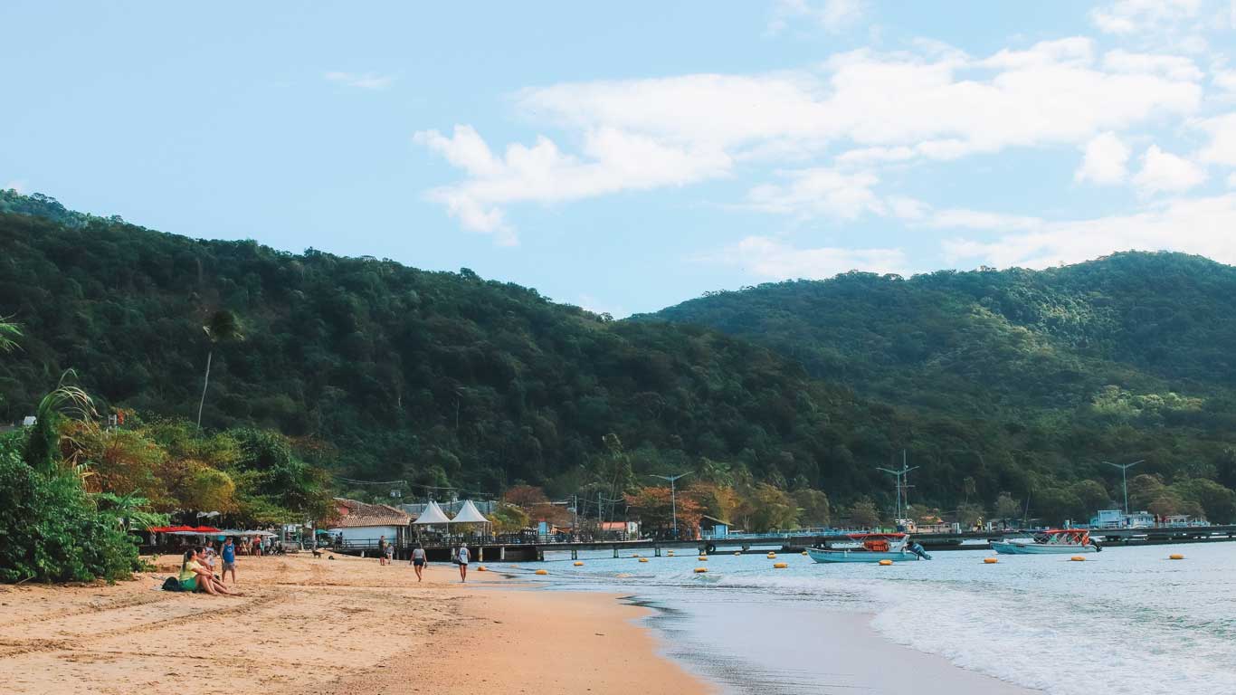 La pintoresca playa en Ilha Grande, Río de Janeiro, con arena dorada que se curva junto a una orilla tranquila. Se ven visitantes relajándose en la arena y caminando cerca del agua, mientras colinas cubiertas de vegetación verde se elevan en el fondo bajo un cielo azul brillante.