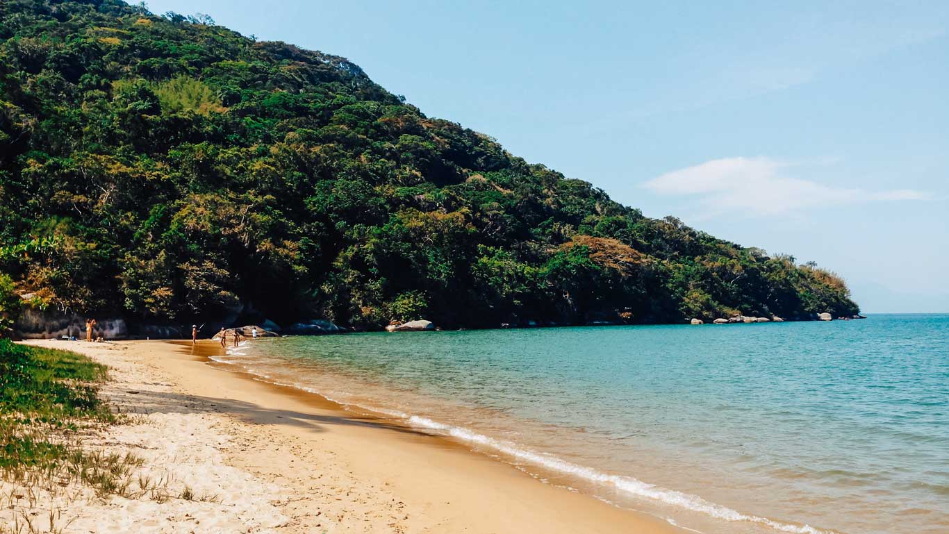 Vista de la Playa do Pouso, una de las playas más tranquilas donde alojarse en Ilha Grande. Esta playa, ubicada en Ilha Grande, Río de Janeiro, se caracteriza por sus arenas suaves y claras que se funden con aguas azules cristalinas. Un denso bosque verde cubre las colinas a la izquierda, mientras algunas personas pasean o se relajan cerca del agua en un día soleado.