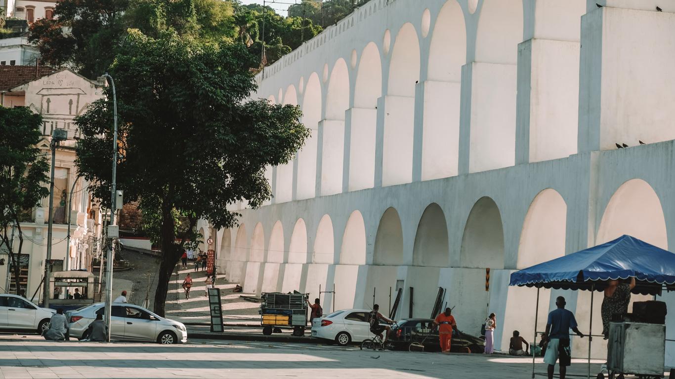 Imagen de los Arcos de Lapa desde otra perspectiva, mostrando la larga fila de arcos blancos que contrastan con el cielo y la vegetación. Se observa la actividad cotidiana con personas caminando, autos estacionados y un pequeño puesto con un toldo azul. En el fondo, las calles empedradas y la arquitectura colonial reflejan la historia del barrio de Lapa.