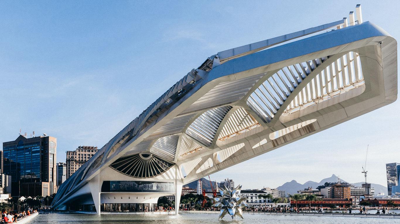 Vista exterior del futurista Museo del Mañana, con su estructura blanca y angular que parece flotar sobre el agua, ubicado en la zona portuaria de Río de Janeiro. Al fondo se aprecian edificios modernos y montañas bajo un cielo despejado.