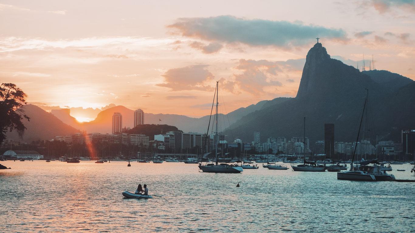 Atardecer sobre la Bahía de Guanabara con varias embarcaciones flotando suavemente. Al fondo, se distingue el icónico Cristo Redentor sobre el cerro del Corcovado y edificios de la ciudad iluminados por la cálida luz del sol poniente.
