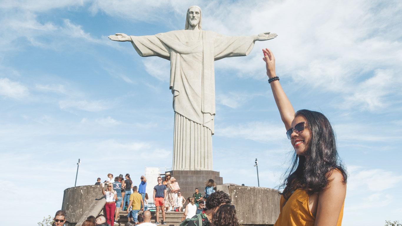 Turista sonriente posa levantando el brazo como si tocara la mano del Cristo Redentor. Alrededor, otros visitantes disfrutan del famoso mirador en un día soleado con cielo azul.